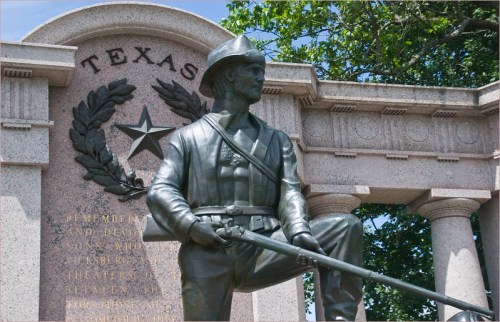A bronze statue of a soldier stands in front of a monument with the word "Texas" and decorative elements.