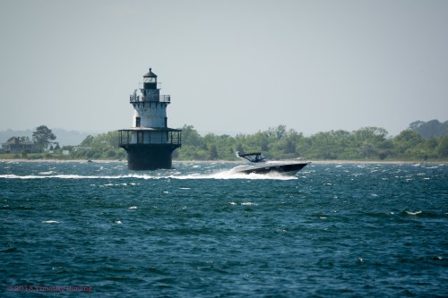 A boat speeds past a tall lighthouse on a sunny day, with waves gently rippling in the foreground.
