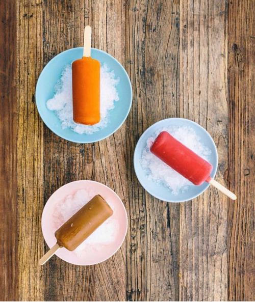 Three colorful popsicles on plates filled with ice, arranged on a wooden table.