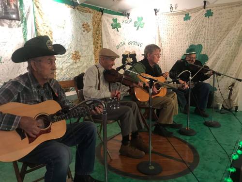 Four musicians perform on stage, playing guitar and fiddle, with a festive backdrop of green decorations.