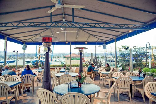 Outdoor dining area with tables and chairs, heaters, and a view of boats in the marina.