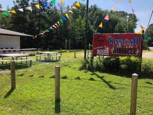Outdoor barbecue restaurant with colorful flags, picnic tables, and a sign reading "Kingfish Barbecue." Green grass surrounds the area.