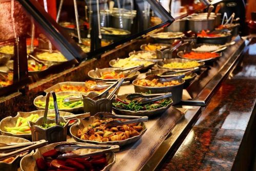 A buffet spread featuring various dishes in decorative bowls, with tongs for serving, set on a polished countertop.