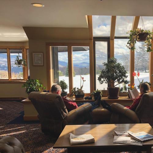 Two people relax in a cozy room with large windows, overlooking a snowy landscape and mountains in the distance.