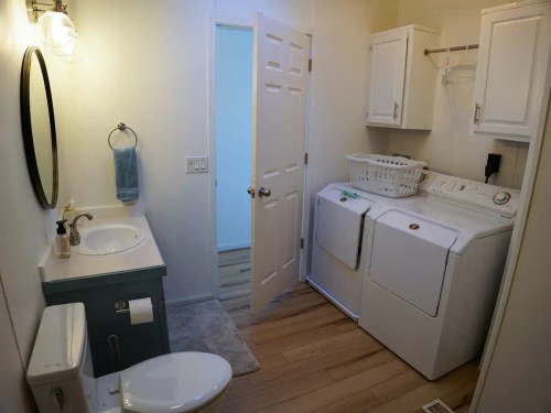 A bright laundry room with a washer, dryer, sink, and a door leading to another room. Light wood flooring.
