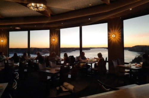 A restaurant interior with diners enjoying a sunset view over a river through large windows.