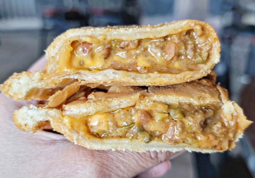 A hand holding a cross-section of a golden-brown stuffed pastry filled with meat and melted cheese.