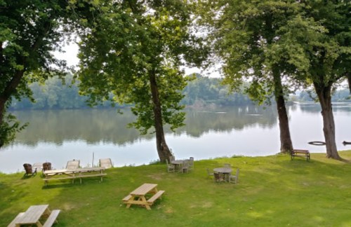 A serene riverside scene with picnic tables and lush green trees reflecting in calm water.
