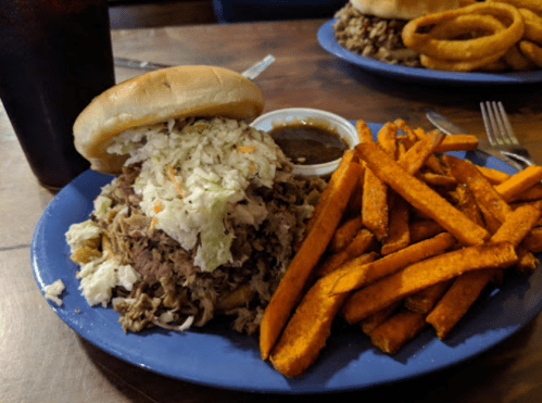 A plate featuring a pulled pork sandwich topped with coleslaw, sweet potato fries, and a side of sauce.