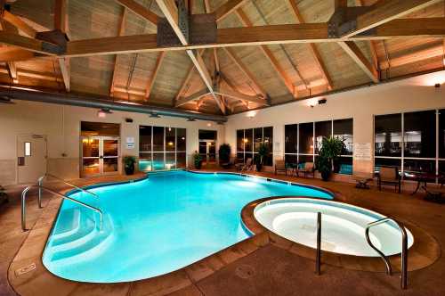 Indoor swimming pool with a hot tub, surrounded by large windows and wooden beams, illuminated with soft lighting.