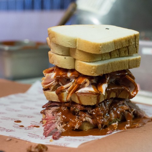 A towering sandwich with layers of meat and barbecue sauce, stacked between slices of white bread on a paper-lined surface.