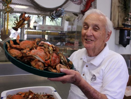 An elderly man smiles while holding a large platter of crabs in a seafood market.
