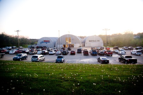 A parking lot filled with cars in front of a large, curved building at sunset, surrounded by green grass and dandelions.