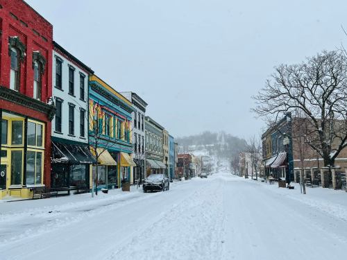 Snow-covered street lined with colorful buildings, shops, and trees, creating a serene winter scene.