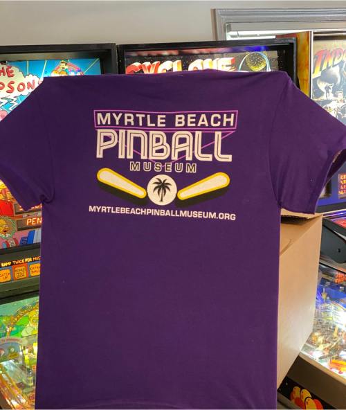 Purple t-shirt featuring the Myrtle Beach Pinball Museum logo and website, displayed in front of pinball machines.