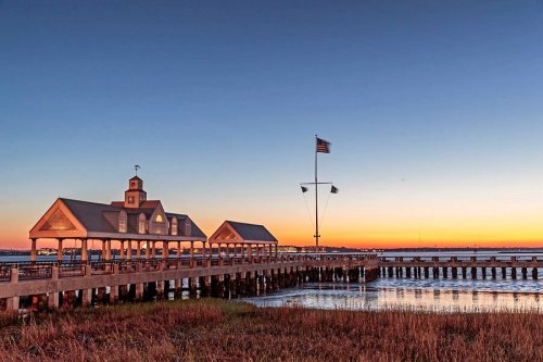A scenic pier at sunset, with an American flag and calm waters reflecting the colorful sky.