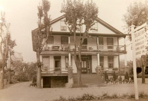 Historic two-story house with balconies, surrounded by trees and a sign indicating a drive-in. Black and white photo.