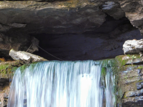A cascading waterfall flows over rocky ledges, surrounded by moss and stone under a dark cave-like overhang.