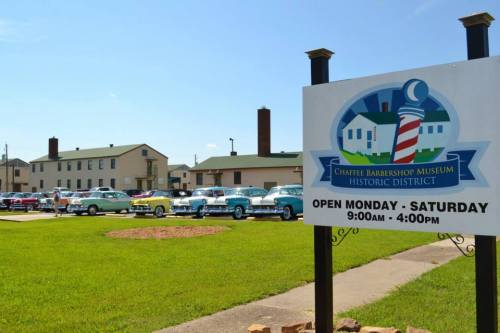 A sign for the Chaffee Barbershop Museum with classic cars lined up in the historic district on a sunny day.
