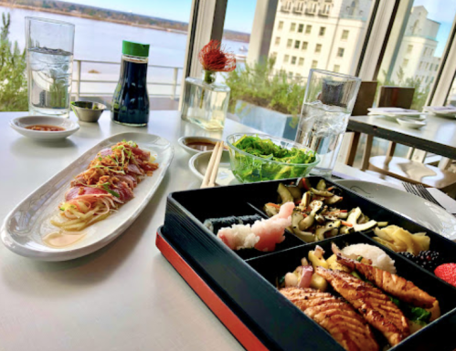 A beautifully arranged sushi meal with sashimi, a bento box, and a view of the water in a modern restaurant setting.