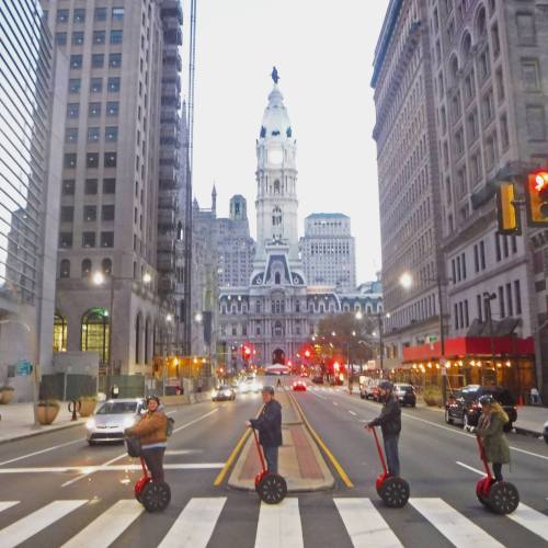 A group of people on Segways crossing a street with a historic building in the background, under a cloudy sky.