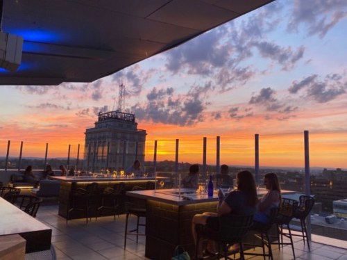 Rooftop view at sunset with people dining, city skyline, and colorful clouds in the sky.