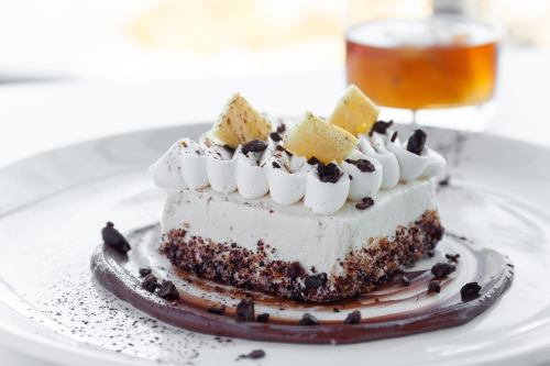 A slice of creamy dessert topped with whipped cream, cake pieces, and chocolate crumbs, served on a plate with a drink.