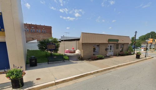 A small ice cream shop with a brown roof, surrounded by greenery and a sidewalk, under a blue sky with clouds.