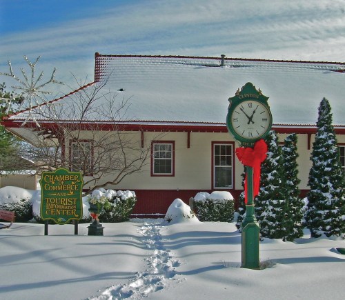 A snowy scene featuring a clock and a sign for a tourist information center, with evergreen trees in the background.