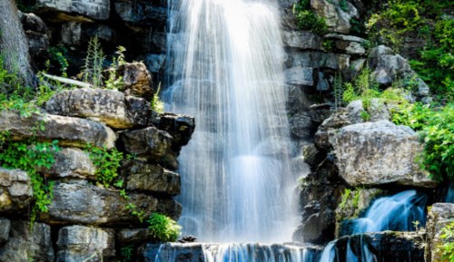 A serene waterfall cascading over rocky cliffs, surrounded by lush greenery and sunlight.