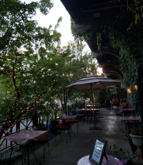 Outdoor dining area with tables under umbrellas, surrounded by greenery and soft lighting.