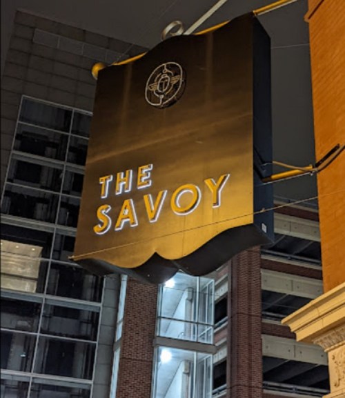 Sign for "The Savoy" illuminated at night, hanging from a building with a modern urban backdrop.