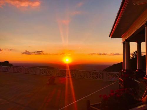 A vibrant sunset casts warm colors over a scenic landscape, with a porch and flowers in the foreground.
