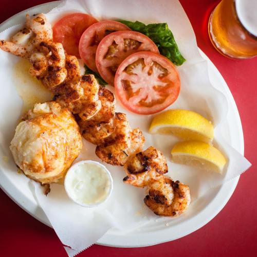 A plate of grilled shrimp skewers, sliced tomatoes, lettuce, lemon wedges, and a side of dipping sauce.
