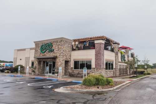 Exterior view of a restaurant with stone accents, outdoor seating, and a cloudy sky. Parking spaces are visible in front.