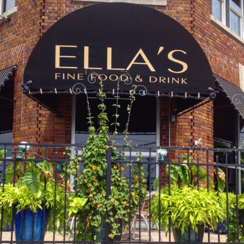 A brick building with a black awning reading "Ella's Fine Food & Drink," surrounded by green plants and a fence.