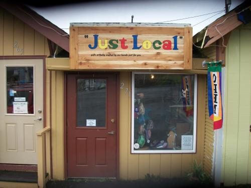 A small storefront with a wooden sign reading "Just Local," featuring a colorful flag and a display of local crafts.