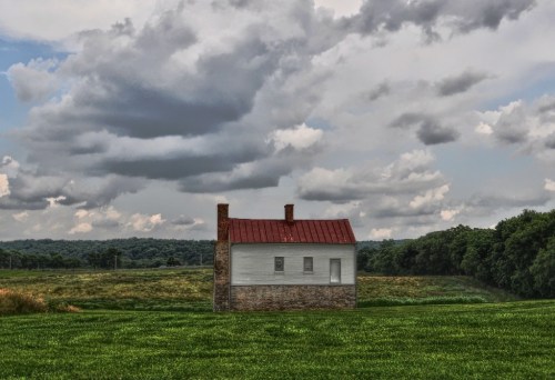 A solitary white house with a red roof stands in a green field under a cloudy sky.
