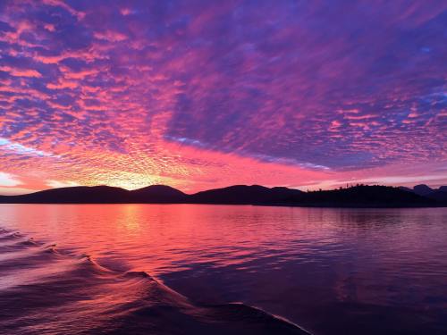 A vibrant sunset over calm waters, with colorful clouds reflecting on the surface and mountains in the background.