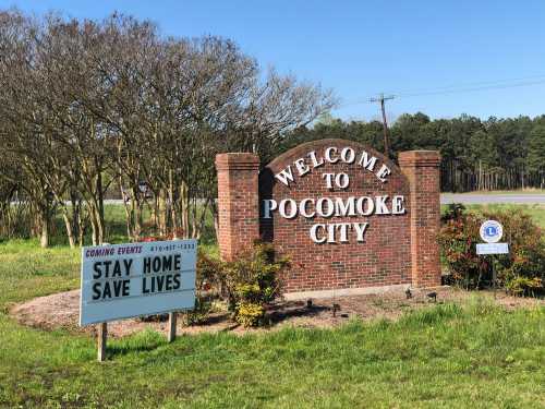 Welcome sign for Pocomoke City with a message to stay home and save lives. Green grass and trees in the background.