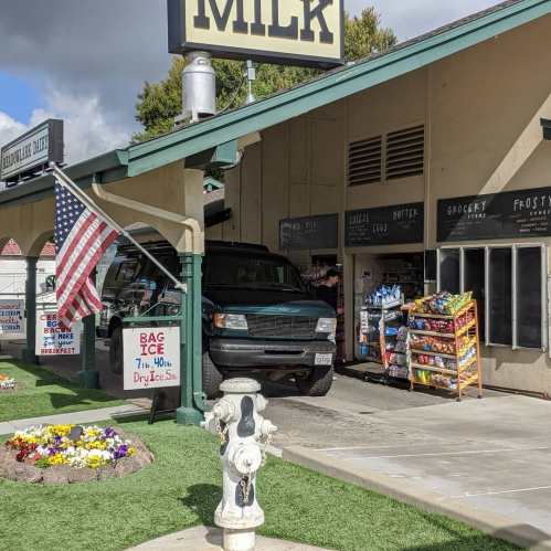 A green truck is parked at a store with an American flag, surrounded by flowers and a display of snacks and drinks.