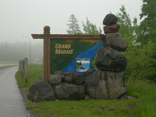 Sign for Grand Marais with stacked rocks, surrounded by greenery and foggy weather, near a road.