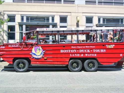 A red amphibious tour bus labeled "Boston Duck Tours" parked near a building, with passengers onboard.