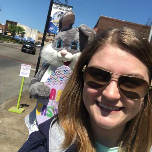 A young woman takes a selfie with a large bunny mascot holding a sign, outdoors on a sunny day.