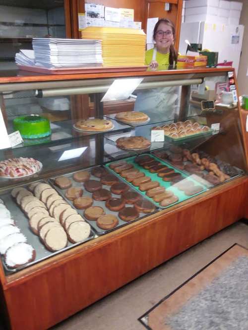 A smiling employee stands behind a glass display case filled with various baked goods and desserts.