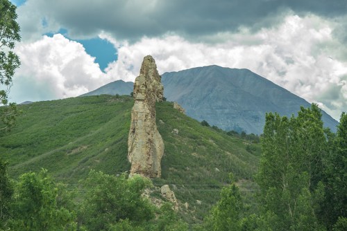 A tall rock formation stands on a green hillside, with a mountain and cloudy sky in the background.