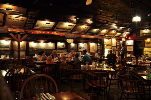 Cozy restaurant interior with wooden tables, patrons dining, and walls adorned with framed artwork and vintage decor.