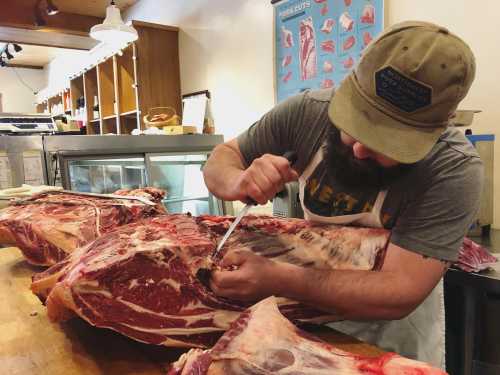 A butcher skillfully cuts meat from a large carcass on a wooden table in a shop.