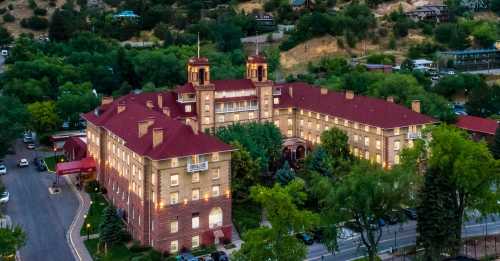 Aerial view of a historic hotel with red roofs, surrounded by trees and hills, illuminated at dusk.