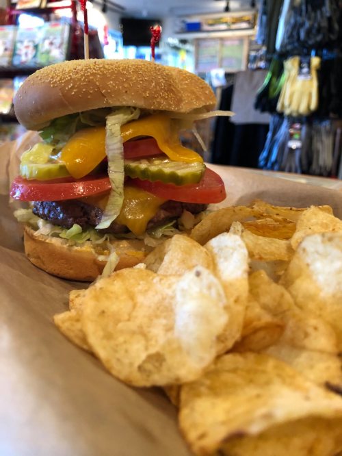 A close-up of a cheeseburger with lettuce, tomato, pickles, and cheese, served with a side of potato chips.